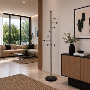 Pode incluir: Um cabideiro moderno com um poste prateado e botões pretos. O cabideiro assenta numa base circular preta. O fundo mostra uma sala de estar com um sofá bege, uma mesa de centro preta e uma grande janela.