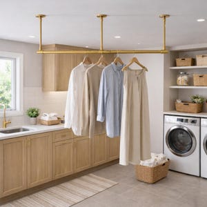Pu&ograve; includere: Un appendiabiti in metallo dorato montato a soffitto sopra un piano cucina. Quattro grucce in legno sostengono una camicia bianca, un top beige a maniche lunghe, una camicia a righe blu e bianche e un lungo abito beige senza maniche.