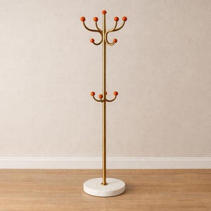 Puede incluir: Un perchero de pie con un poste central de metal dorado, una base circular de mármol blanco y dos niveles de ganchos metálicos curvos. Cada gancho está rematado con una pequeña bola redonda de color naranja brillante y contrastante.