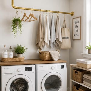 May include: A gold-toned metal pipe clothing rack mounted to the ceiling and wall, featuring S-hooks holding wooden hangers, hanging towels, and a beige laundry tote bag. A framed sign on the wall reads WASH DRY FOLD REPEAT.