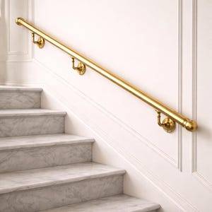 Puede incluir: Una barandilla de latón pulido está montada en una pared blanca junto a una escalera de mármol. La barandilla está soportada por soportes decorativos y tiene un extremo redondeado. La escalera tiene escalones de mármol gris y blanco.