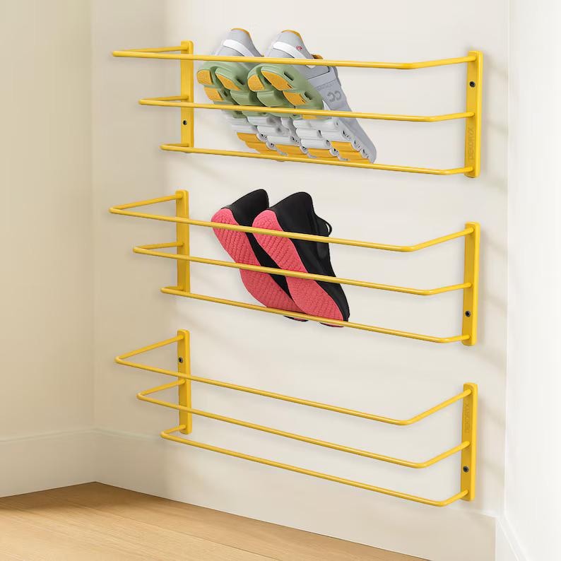 May include: Three yellow metal wall-mounted shoe racks with horizontal bars. The top rack holds a pair of grey and light green athletic trainers. The middle rack holds a pair of black athletic trainers with pink soles. The bottom rack is empty.
