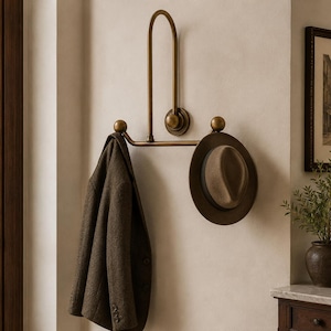 Puede incluir: Un perchero de pared de latón con un marco vertical en forma de U y dos brazos horizontales que terminan en pomos esféricos. Un blazer de lana marrón oscuro cuelga del lado izquierdo y un sombrero fedora de fieltro marrón cuelga del derecho.