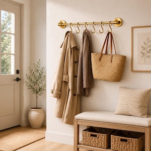Op de afbeelding: Een goudkleurige metalen wandkapstok met een horizontale stang en zeven S-vormige haken. Aan de haken hangen een beige trenchcoat, een bruine sjaal en een gevlochten strooien tas met bruine lederen handvatten.