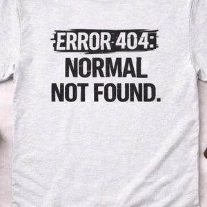 Op de afbeelding: Lichtgrijs t-shirt met de zwarte tekst "ERROR 404: NORMAL NOT FOUND". Er omheen liggen een zwarte koptelefoon, zonnebril, horloge, telefoon, cameralens en zwart-witte sneakers.