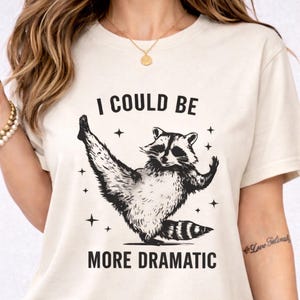 Puede incluir: Camiseta color crema con un gráfico de mapache en blanco y negro en una pose dramática, rodeado de estrellas. El texto "I COULD BE MORE DRAMATIC" está impreso en negro, añadiendo un toque de humor al diseño.