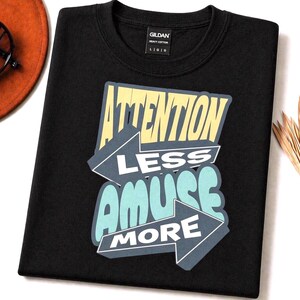 Op de afbeelding: Zwart t-shirt met de tekst "ATTENTION LESS AMUSE MORE" in geel, blauw en wit. De afbeelding is ontworpen met pijlaccenten. Het shirt is gemaakt van zwaar katoen.