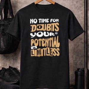 Puede incluir: Camiseta negra con el texto "NO TIME FOR DOUBTS YOUR POTENTIAL IS LIMITLESS" en letras blancas y doradas. La camiseta está colgada de una percha de madera. Otros artículos en el fondo incluyen una bolsa de deporte negra, zapatos y una botella de agua.