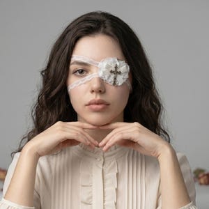 Può includere: Una benda per occhio in pizzo bianco caratterizzata da una rosetta in tessuto arricciato e un ciondolo a forma di croce metallica centrale. La benda è fissata con sottili lacci in pizzo bianco che avvolgono la testa, offrendo una copertura decorativa.