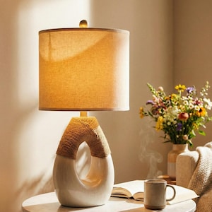 May include: A white ceramic table lamp with a beige fabric shade. The lamp base has a unique design with a rope-wrapped section. A small, round, clear finial tops the lamp. The lamp is on a round table with a book, mug, and vase of flowers.