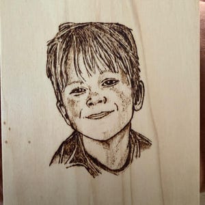 Pode incluir: Retrato pirograbado de uma criança sorrindo em uma placa de madeira. A obra de arte apresenta uma representação detalhada do rosto e cabelo da criança. Uma corda escura está presa para pendurar a placa.