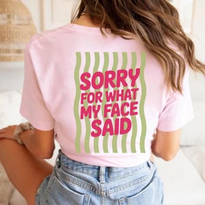 Könnte beinhalten: Hellrosa T-Shirt mit dem Text "SORRY FOR WHAT MY FACE SAID" in leuchtendem Pink, umrahmt von wellenförmigen grünen Streifen. Ein lässiges Kleidungsstück.