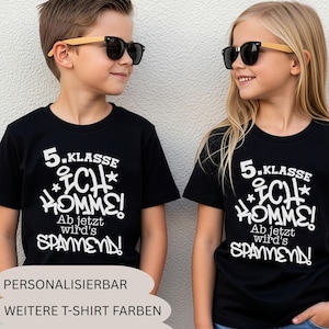 Op de afbeelding: Twee zwarte t-shirts met korte mouwen en de witte grafische tekst 5. KLASSE ICH KOMME! Ab jetzt wird's SPANNEND! De t-shirts worden gedragen door twee kinderen met een zonnebril met lichtgekleurde houten monturen tegen een muur.