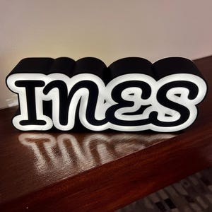 Può includere: Un'insegna luminosa in bianco e nero con il nome "INES". Le lettere spesse hanno un contorno nero e un interno bianco brillante. L'insegna è posizionata su una superficie di legno scuro, creando un effetto visivo sorprendente.