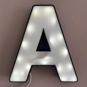 Può includere: Una grande lettera "A" illuminata con un bordo nero. L'interno della lettera è bianco e illuminato da piccole luci dai toni caldi. Un triangolo nero riempie il centro della lettera. Un cavo di alimentazione bianco si estende dal basso.