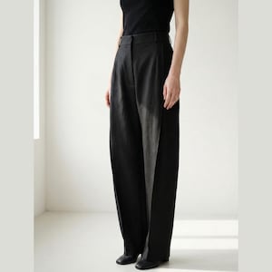 Puede incluir: Pantalones negros de pernera ancha, cintura alta y corte entallado. Los pantalones son de tejido oscuro y presentan un diseño clásico. Los pantalones se combinan con un top negro y zapatos negros.
