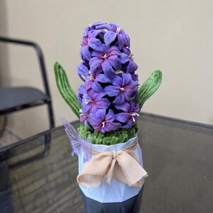 Pode incluir: Um jacinto roxo feito de feltro, com folhas verdes e estames brancos. A flor está num vaso geométrico branco adornado com uma fita de tecido bege e um pequeno detalhe de borboleta roxa translúcida fixado na lateral.