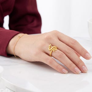 Könnte beinhalten: Ein goldfarbener Ring in Form einer gewundenen Schlange mit strukturierter Oberfläche und kleinen dunklen Augen, der an einem Finger getragen wird. Ein dünnes goldfarbenes Kettenarmband mit einem kleinen dunklen Akzent ist ebenfalls am Handgelenk sichtbar.