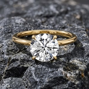 May include: A gold ring with a large, round, clear gemstone. The ring is set on a gray, textured stone surface. The band is a simple, polished gold. The gemstone is held in place by four prongs.