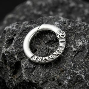 May include: A silver-colored, hinged ring with engraved Tibetan script and a decorative symbol. The ring is circular and appears to be made of metal. The ring is resting on a dark, textured stone surface.