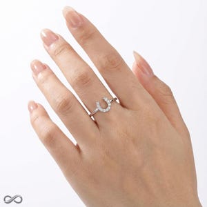 Op de afbeelding: Een zilveren hoefijzervormige ring met een rij kleine, heldere edelstenen. De ring zit om een vinger, met de hand en nagels op de achtergrond.