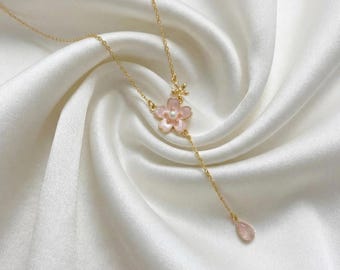 May include: A delicate gold-tone necklace featuring a pink flower pendant with a pearl center and a teardrop-shaped charm. The necklace is displayed on a white satin background, showcasing its elegant design.
