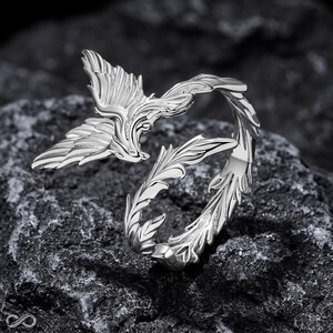 May include: A silver-toned ring featuring an open design with a detailed wing motif on one side and a leafy, vine-like pattern extending along the band. The metal has a polished, reflective finish that highlights the intricate textures of the feathers.