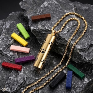 Op de afbeelding: Een goudkleurige cilindervormige hanger aan een bijpassende schakelketting, voorzien van zwarte uitsparingen in de vorm van pootafdrukken. De hanger wordt vergezeld door diverse kleine, rechthoekige viltjes in kleuren zoals rood, groen en paars.