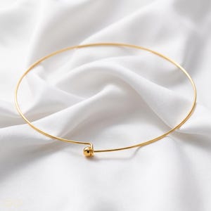 Puede incluir: Un collar tipo gargantilla circular y fino de metal en tono dorado, que presenta una cuenta esférica pulida de color dorado en la parte frontal, donde se unen los dos extremos del alambre. El collar tiene un diseño liso y sencillo.