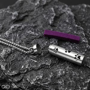 May include: A silver-coloured cylindrical pendant necklace featuring cut-out paw print designs. The pendant is open, revealing a hollow interior, and is accompanied by a small, rectangular purple felt insert. The metal chain is attached to the cap.