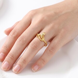 May include: A gold-toned ring shaped like a coiled snake with a textured, scale-like surface. The snake's head is positioned at the top, and its body wraps around the finger to form the band of the ring, featuring a metallic finish.