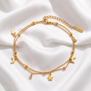 Op de afbeelding: Een goudkleurig enkelbandje met dubbele ketting, voorzien van bungelende ster- en maansikkelbedels, kleine ronde kraaltjes en een helder kristallen accent. De ketting heeft een verstelbaar verlengstuk met een label met S.STEEL.