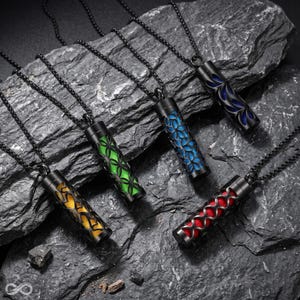 May include: Four cylindrical pendant necklaces with black metal casings featuring intricate cut-out patterns. Each pendant reveals a vibrant inner core in yellow, green, blue, or red, suspended from matching black metal ball chains.