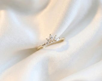 May include: A delicate gold ring featuring a crown design. The ring is adorned with several clear, marquise-cut gemstones. The ring is set against a white, silky background, highlighting its elegant design. A piece of jewelry suitable for special occasions.