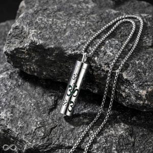 May include: A silver-toned cylindrical pendant necklace featuring a repeating Om symbol cutout design. The pendant is attached to a matching silver-toned ball chain, creating a cohesive metallic look with a polished finish and intricate detailing.
