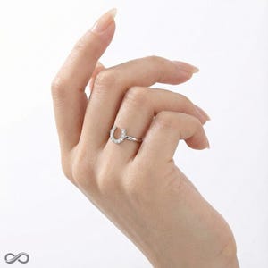 Op de afbeelding: Een zilveren hoefijzervormige ring met kleine, heldere edelstenen langs de boog. De ring heeft een smalle band en wordt op een hand getoond met een lichte achtergrond.