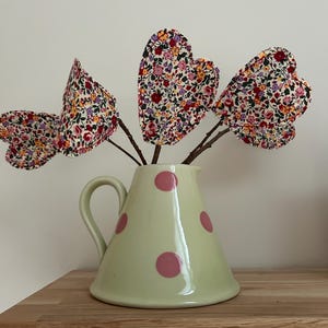 May include: A light green ceramic pitcher with pink polka dots holds heart-shaped fabric flowers. The flowers have a floral pattern with red, pink, and yellow hues. The pitcher sits on a wooden shelf.