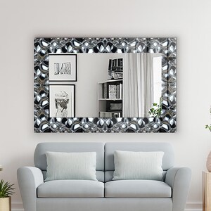 May include: A rectangular mirror with a decorative, textured frame in shades of silver and black hangs above a light blue sofa. The mirror reflects a room with artwork, a bookshelf, and a window with white curtains. The room has a light wood floor.