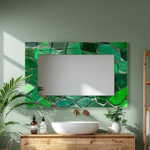 May include: A rectangular mirror with a mosaic-style frame in shades of green. The frame is composed of irregularly shaped pieces, creating a textured appearance. The mirror is mounted on a light green wall, above a wooden vanity with a white sink.