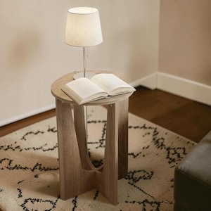 May include: A small, round wooden side table with a lamp and an open book. The lamp has a beige lampshade and a silver base. The table is light brown and has a unique leg design. The open book has white pages.