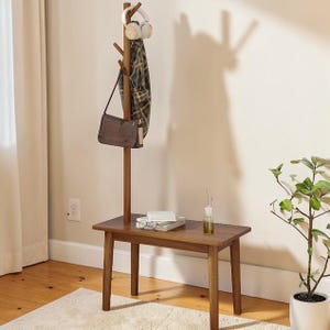 May include: A wooden coat rack with a small bench. A brown purse, plaid scarf, and headphones hang from the rack. A stack of books and a small vase sit on the bench. A potted plant is in the corner.