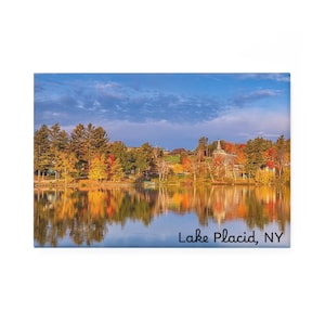 Puede incluir: Un imán rectangular con una vista panorámica de Lake Placid, NY. La imagen muestra un lago que refleja árboles otoñales coloridos bajo un cielo azul. El texto "Lake Placid, NY" está escrito en una fuente cursiva.