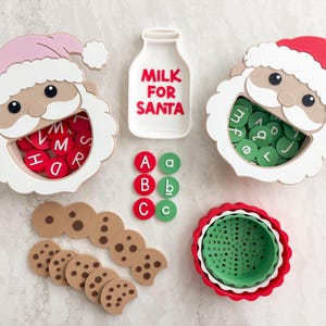 May include: Two Santa-shaped containers with white beards, one wearing a pink hat and one wearing a red hat, filled with red and green letter tokens. Includes a milk bottle-shaped tray labeled MILK FOR SANTA, cookie-shaped tokens, and a green bowl.