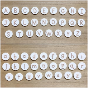 May include: A collection of white circular discs, each featuring a single raised letter in a light brown color. The set includes both uppercase and lowercase letters of the English alphabet, arranged in rows. Each disc displays a distinct character.