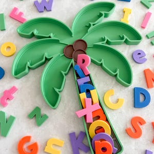 May include: A green palm tree-shaped container with three brown circles at the top, filled with various colorful plastic letters. The container is surrounded by a scattered collection of individual plastic letters in multiple bright colors.
