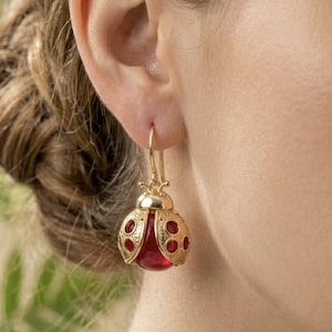 Peut inclure: Boucles d'oreilles coccinelle dorées avec des accents rouges. Les boucles d'oreilles présentent un design détaillé avec un corps rouge arrondi et des ailes en filigrane dorées. Les boucles d'oreilles sont suspendues à un crochet doré.