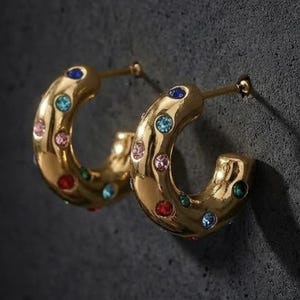 May include: A pair of gold-colored hoop earrings. Each earring is adorned with a series of colorful gemstones, including blue, pink, red, and green. The earrings have a polished finish and are set against a dark background.