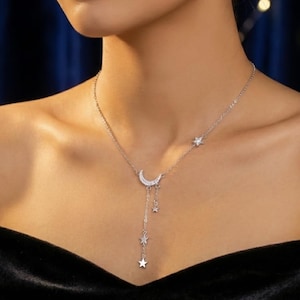Peut inclure: Collier argenté orné d'un pendentif en forme de croissant de lune orné de petites pierres scintillantes. Le collier est doté d'une chaîne délicate avec des breloques en forme d'étoiles pendantes et une étoile sur le côté. Le bijou est porté sur fond sombre.