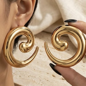 May include: Gold-coloured spiral earrings. One earring is worn on an ear, and another is held by a hand. The earrings have a smooth, polished finish and a curved, swirling design. The earrings are a bold, statement piece.