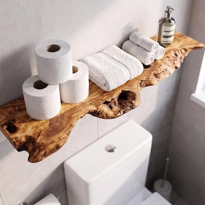 May include: A live edge wooden shelf featuring a natural grain pattern and irregular bark edges. The shelf holds three white toilet paper rolls, a folded white towel, two rolled white hand towels, and a clear glass bottle with a pump dispenser.